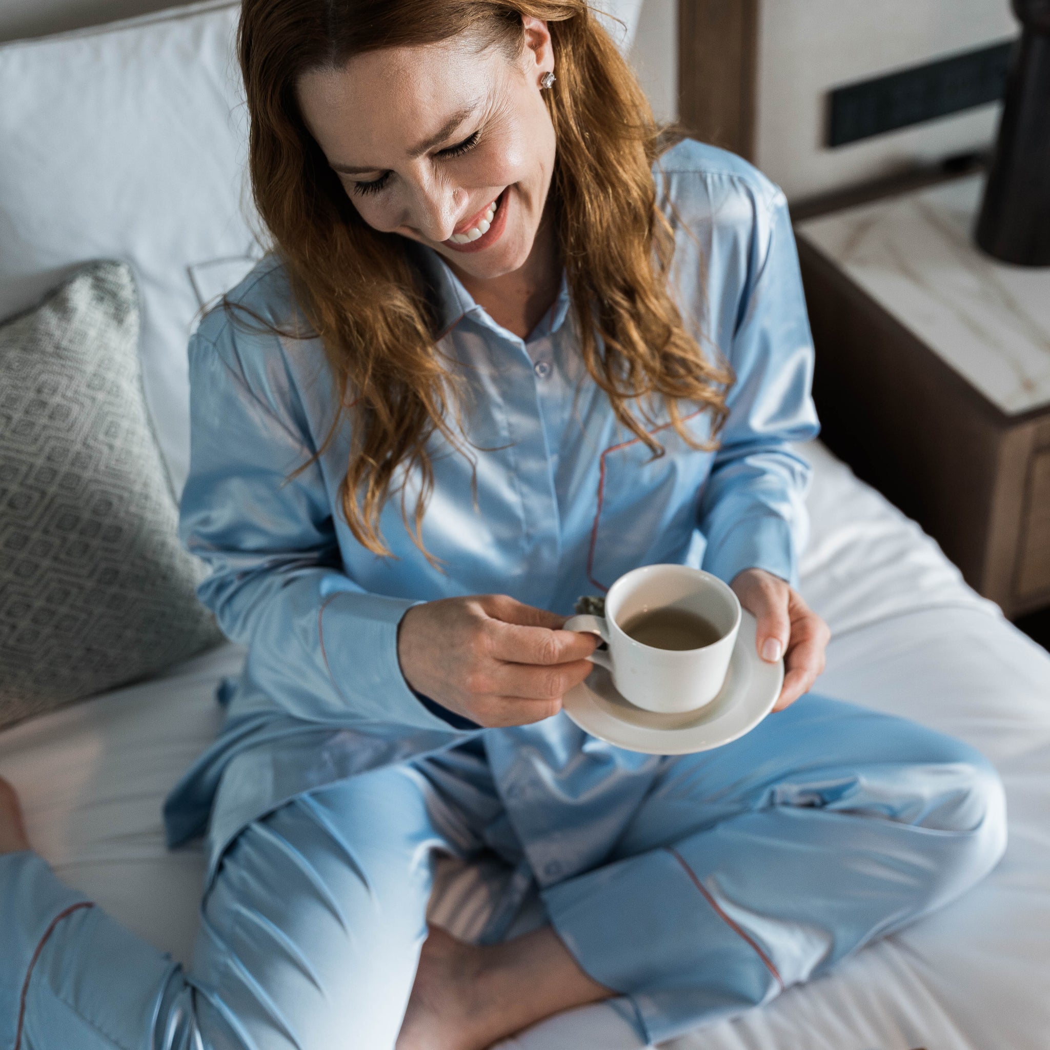 Woman in light blue pajamas sitting on a bed holding a cup of tea.