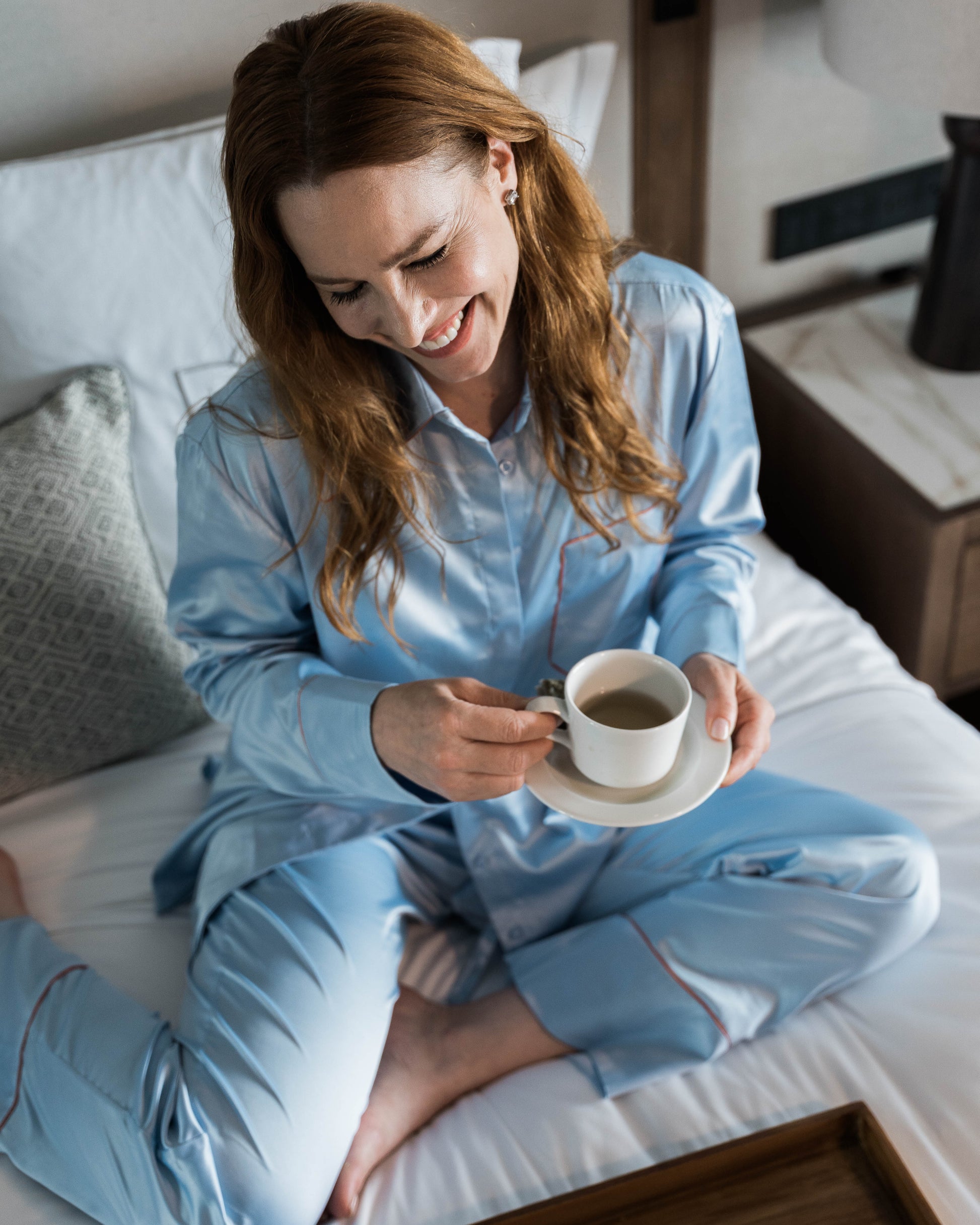 Woman in light blue pajamas sitting on a bed holding a cup of tea.
